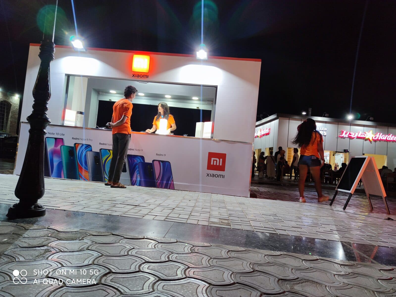 A staffed, branded Xiaomi/Redmi kiosk with a full product lineup poster at a seaside retail strip at night